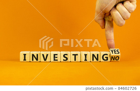 Investing yes or no symbol. Businessman turns a wooden cube and change words 'investing no' to 'investing yes'. Beautiful orange background. Business and investing yes or no concept, copy space. Investing yes or no symbol. Businessman turns a wooden cube and change words 'investing no' to 'investing yes'. Beautiful orange background. Business and investing yes or no concept, copy space. 84602726