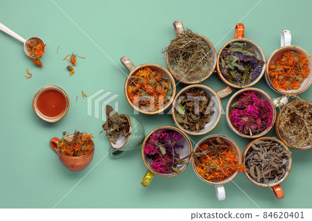 Assortment of dried relaxing tea herbs in colourful cups on mint green background with honey. Calendula, mint, anise hyssop, monarda didyma, wormwood, sage leaves. 84620401