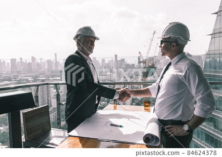 Two engineering working together in their new project. They are wearing suit , tie and white hard hat. They are looking at their design on the paper on the wooden table Two engineering working together in their new project. They are wearing suit , tie and white hard hat. They are looking at their design on the paper on the wooden table 84624378