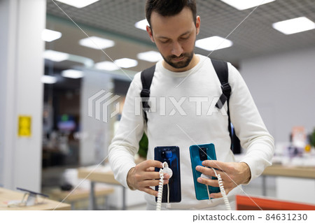 Photo of an adult handsome thinking brunette man with stubble in a white sweatshirt chooses a mobile 84631230