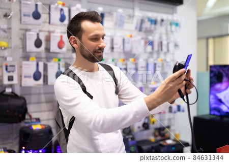 Photo of an adult handsome thinking brunet man with stubble in a white sweatshirt chooses a camera Photo of an adult handsome thinking brunet man with stubble in a white sweatshirt chooses a camera 84631344