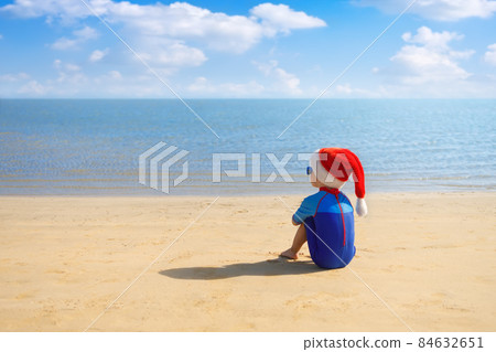 back view of child boy in santa hat, sunglasses and swimsuit sitting on the sand beach back view of child boy in santa hat, sunglasses and swimsuit sitting on the sand beach 84632651