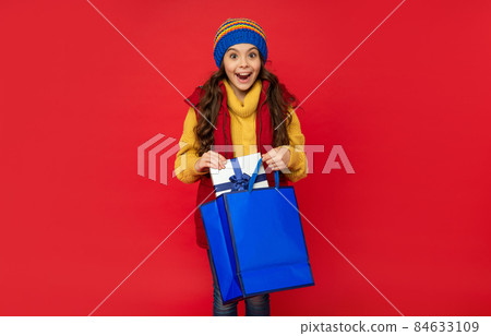 amazed teen girl wearing knitted hat hold shopping bag and present box on red background, purchase amazed teen girl wearing knitted hat hold shopping bag and present box on red background, purchase 84633109
