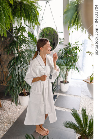 Full length shot of cheerful young woman in white bathrobe smiling while applying lip balm on her lips, standing in the bathroom decorated with plants 84633704