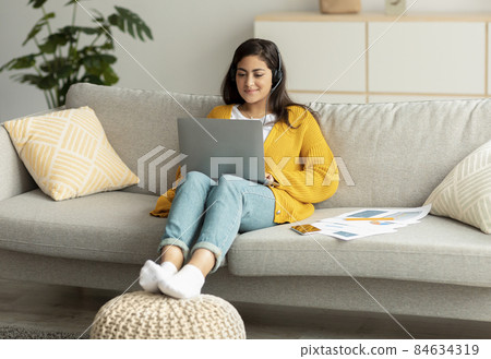 Telecommuting concept. Excited arab woman sitting on sofa and working online on laptop, wearing headset 84634319