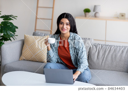 Lovely young Arab lady using laptop, studying or working online, having remote business meeting, drinking coffee at home Lovely young Arab lady using laptop, studying or working online, having remote business meeting, drinking coffee at home 84634403
