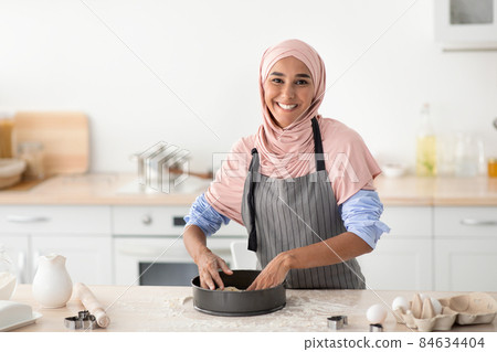 Happy Muslim Woman Pressing Dough Into Mold While Baking In Kitchen Happy Muslim Woman Pressing Dough Into Mold While Baking In Kitchen 84634404