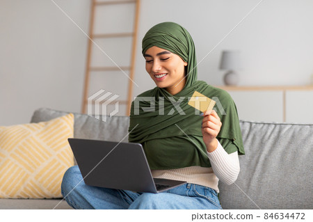 Young Arab female in hijab using credit card and laptop, shopping online on couch, buying goods on internet from home 84634472