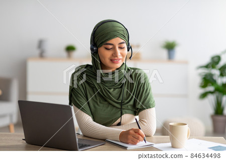 Young Arab woman in hijab working on laptop, wearing headphones, taking notes during webinar at home 84634498