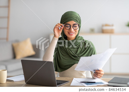 Young Arab female freelancer in hijab working with papers and laptop at home 84634534