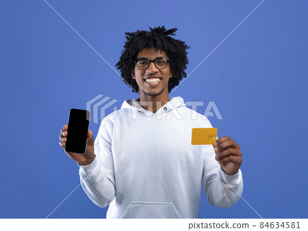 Happy black teenager holding credit card and mobile phone on blue studio background, mockup for app or website Happy black teenager holding credit card and mobile phone on blue studio background, mockup for app or website 84634581