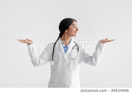 Smiling millennial indian woman doctor in white coat raises her hands and makes choice, isolated on light background Smiling millennial indian woman doctor in white coat raises her hands and makes choice, isolated on light background 84634906