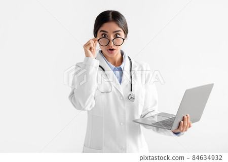 Surprised young hindu female doctor in white coat takes off glasses and looks at camera, works at laptop 84634932