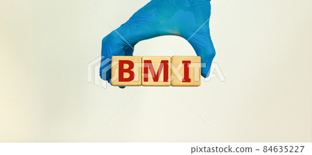 Medical BMI concept. Hand in blue glove holds wooden cubes with the inscription 'BMI - biomedical research or body mass index'. Beautiful white background. Copy space. Medical BMI concept. Hand in blue glove holds wooden cubes with the inscription 'BMI - biomedical research or body mass index'. Beautiful white background. Copy space. 84635227