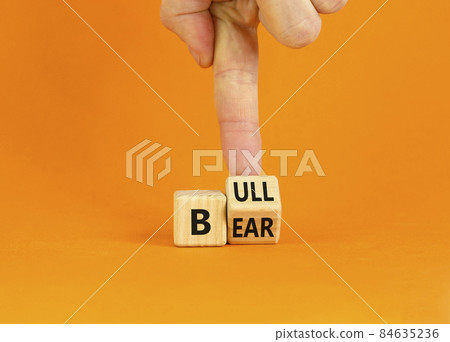 Symbol for a positive trend at the stock market. Businesman turns a wooden cube and changes the word 'bear' to 'bull'. Beautiful orange background. Business and bear or bull concept. Copy space. Symbol for a positive trend at the stock market. Businesman turns a wooden cube and changes the word 'bear' to 'bull'. Beautiful orange background. Business and bear or bull concept. Copy space. 84635236