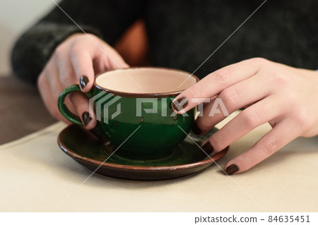 young female hands with dark nail polish holding beautiful dark green coffee cup on platter with hot green tea young female hands with dark nail polish holding beautiful dark green coffee cup on platter with hot green tea 84635451