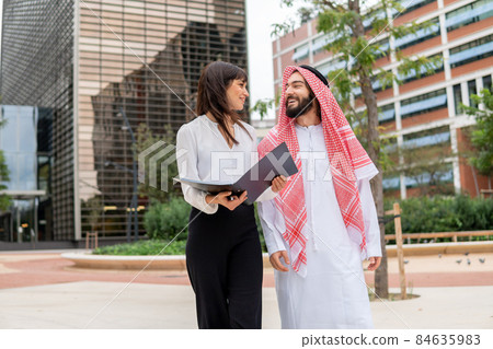 Businesswoman with folder looking at happy Arab man while walking on city street during business meeting together Businesswoman with folder looking at happy Arab man while walking on city street during business meeting together 84635983
