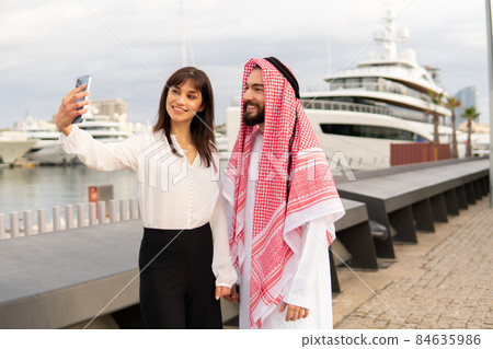 Female entrepreneur smiling and taking selfie with Arab man in traditional clothes while standing on embankment near luxury ship together Female entrepreneur smiling and taking selfie with Arab man in traditional clothes while standing on embankment near luxury ship together 84635986
