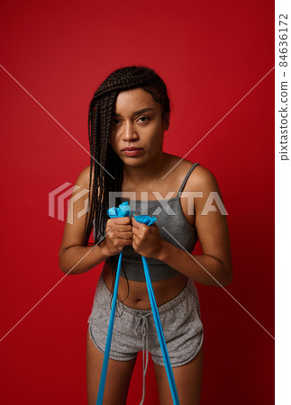Strong African American woman with stylish dreadlocks using a resistance band in her exercise routine. Female athlete exercising with resistance band against red colored background with copy space 84636172