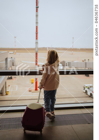 a little girl with a suitcase stands by the window at the airport.  84636738