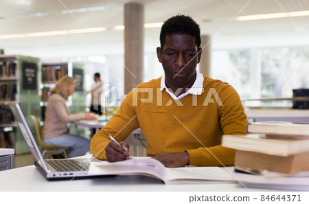 Efficient man preparing for exam in public library, reading book and using laptop for searching information 84644371
