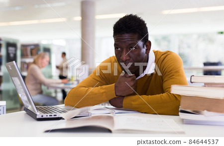 Thoughtful african-american man working with textbook and laptop in library 84644537
