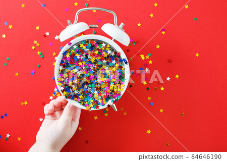 child's hands hold alarm clock with scattered sequins on a red background. christmas, celebration concept 84646190