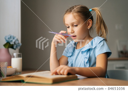Close-up of thinking pretty elementary child school girl doing homework and holding pen against mouth sitting at home table. Close-up of thinking pretty elementary child school girl doing homework and holding pen against mouth sitting at home table. 84651036