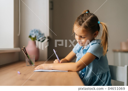 Side view of focused primary little child girl learning writing doing homework sitting at home table by window. Side view of focused primary little child girl learning writing doing homework sitting at home table by window. 84651069