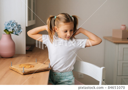 Portrait of tired pretty primary little girl stretching out during doing herbarium sitting at home table by window. 84651070