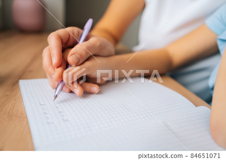 Close-up cropped shot of primary daughter doing homework writing in copybook with pen with young mother sitting at home table. Close-up cropped shot of primary daughter doing homework writing in copybook with pen with young mother sitting at home table. 84651071