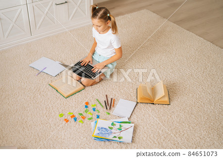 High-angle view of primary little child girl doing homework using laptop computer for distance education online sitting on floor at home. High-angle view of primary little child girl doing homework using laptop computer for distance education online sitting on floor at home. 84651073