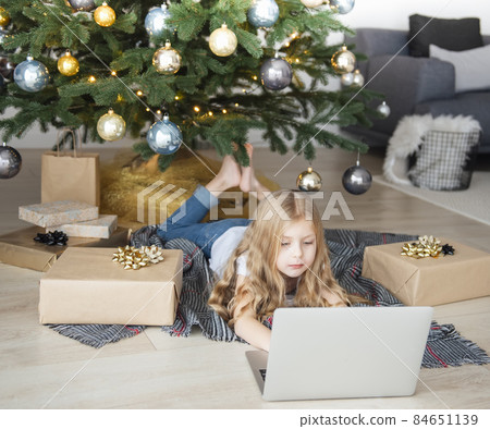 Little girl lies near the Christmas tree with a laptop. 84651139