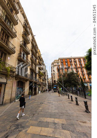 Barcelona, Spain - September 23, 2021: Cityscape of Barcelona. Typical architecture in the Catalan capital. Narrow streets and houses with narrow iron grate balconies. People walk on the streets. Barcelona, Spain - September 23, 2021: Cityscape of Barcelona. Typical architecture in the Catalan capital. Narrow streets and houses with narrow iron grate balconies. People walk on the streets. 84651545