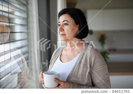 Sad senior woman standing and looking out through window indoors at home. 84651765