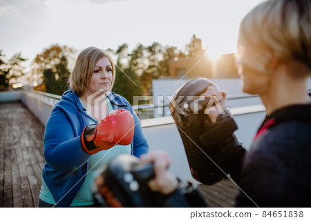 Overweight woman training boxing with personal trainer outdoors on terrace. 84651838