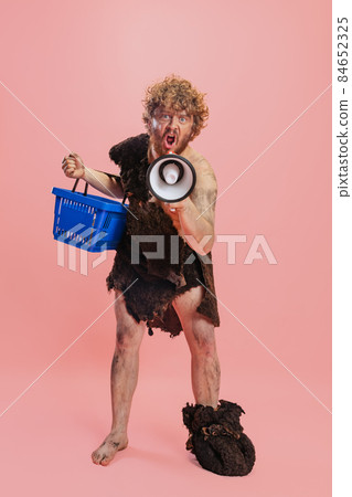 Portrait of man in character of neanderthal, primitive person shouting in megaphone and holding shopping basket isolated over pink background 84652325