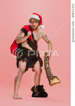 Full-length portrait of man in character neanderthal, primitive person holding log and Christmas presents bag isolated over pink background 84652341