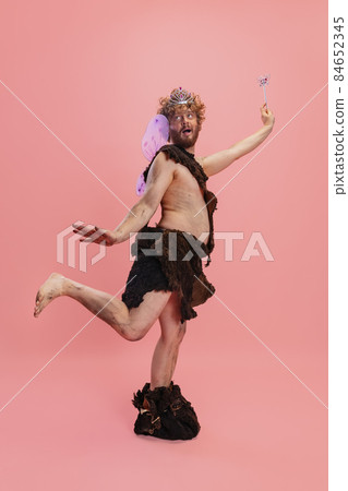 Portrait of man in character of neanderthal wearing fairy wings and crown posing like little wizzard isolated over pink background. CVute 84652345