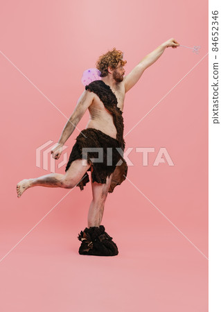 Full-length portrait of man in character of neanderthal wearing fairy wings and crown posing like little wizzard isolated over pink background 84652346