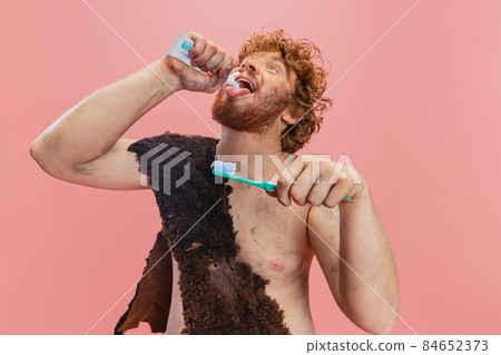 Portrait of man in character of neanderthal squeezing tootpaste into mouth isolated over pink background 84652373