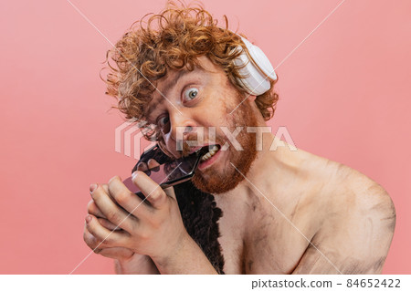 Cropped portrait of man in character of primitive human, neanderthal in headphones biting phone isolated over pink background 84652422