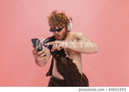 Cropped portrait of man in character of neanderthal using phone and listening to music in headphones isolated over pink background 84652450