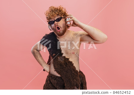 Cropped portrait of man in character of neanderthal wearing stylish sunglasses and listening to music in headphones isolated over pink background Cropped portrait of man in character of neanderthal wearing stylish sunglasses and listening to music in headphones isolated over pink background 84652452