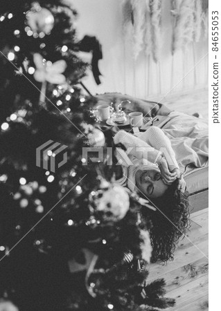 A young woman in cozy home clothes poses on a bed in a loft interior near a Christmas tree. 84655053