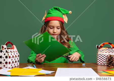 Little cute girl dressed like funny gnome or elf sitting at table and making paper Christmas decorations isolated over green background. 84656673