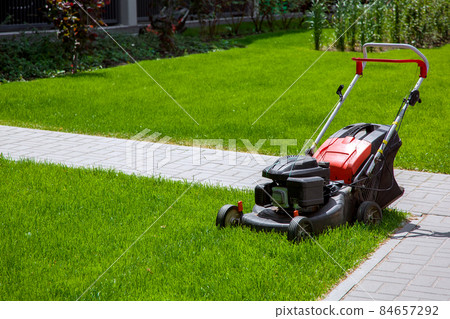gasoline engine lawn mower for landscaping and backyard lawn mowing, gardening equipment on green grass surrounded by stone sidewalk walkway on a sunny summer day front view, nobody. gasoline engine lawn mower for landscaping and backyard lawn mowing, gardening equipment on green grass surrounded by stone sidewalk walkway on a sunny summer day front view, nobody. 84657292