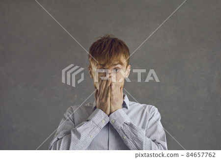 Oops. Close up portrait of attractive man with closed mouth, hands on face and eyes wide open, looking at the camera, he cannot believe his eyes, standing against gray background 84657762