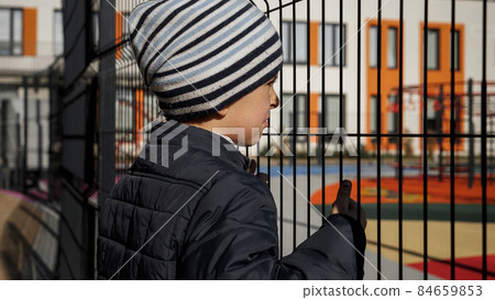 Little upset boy looking through metal fence on modern school and playground. Concept of poverty, immigration, bullying and kids stress 84659853