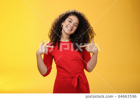 Proud and satisfied ambitious successful female student in red dress standing pleased smiling and pointing at herself as if bragging about own achievements happily and glad over yellow wall 84663596